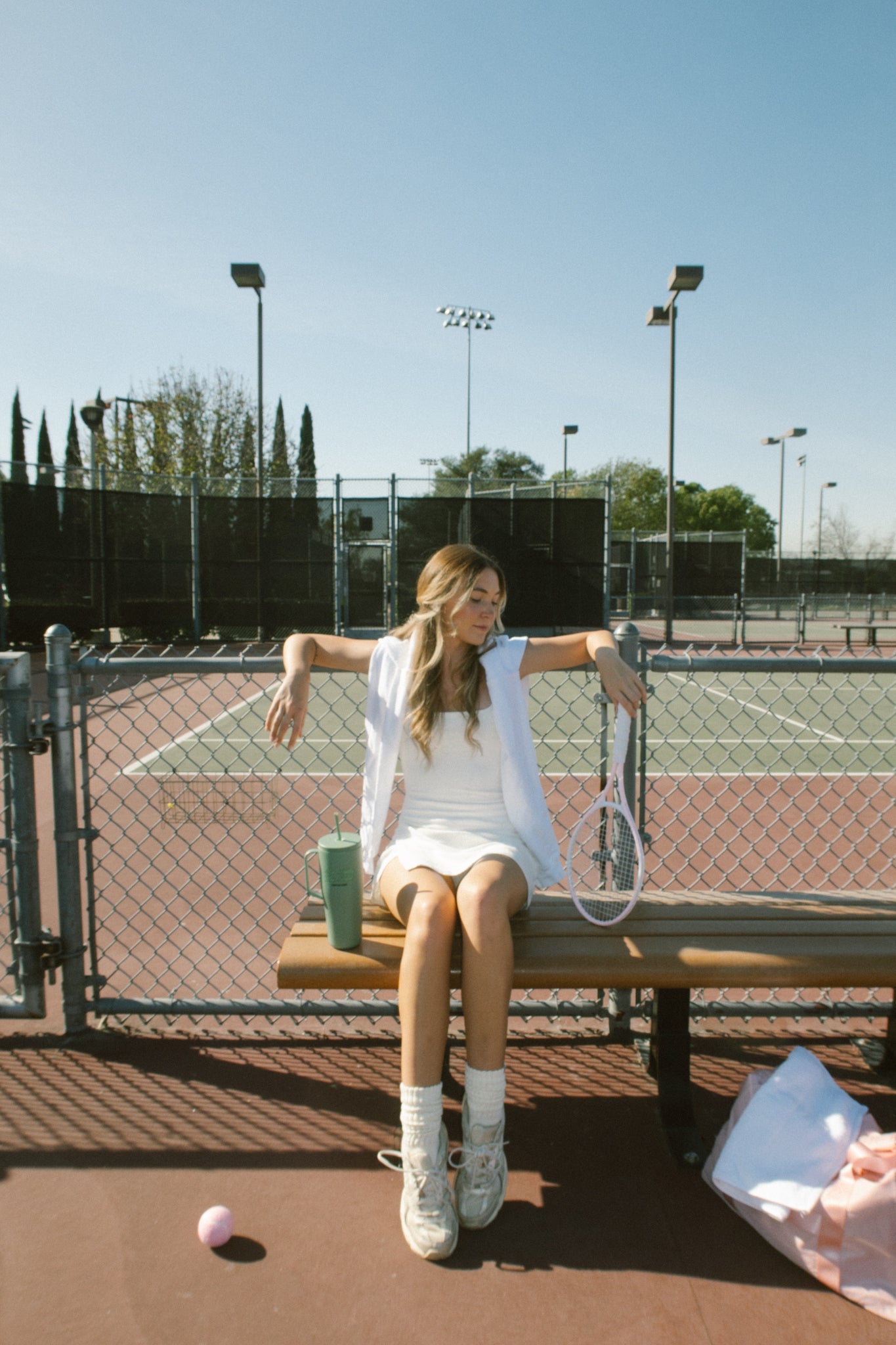 Blonde young woman sitting on a bench on a tennis court with a pink tennis racket and green tumbler with straw that says make today your bitch.