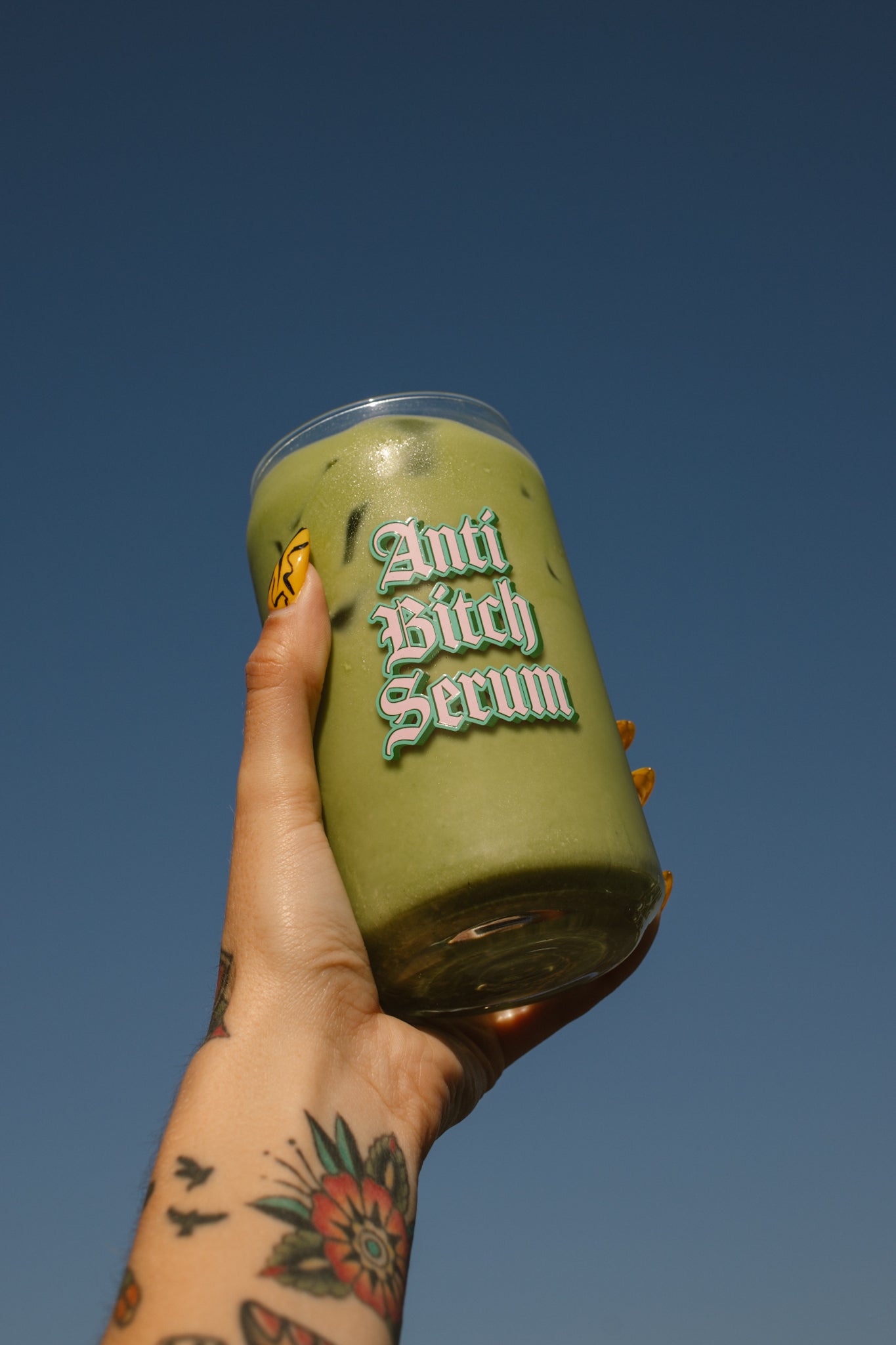 Hand holding a green cup with 'Anti Bitch Serum' text against a clear blue sky
