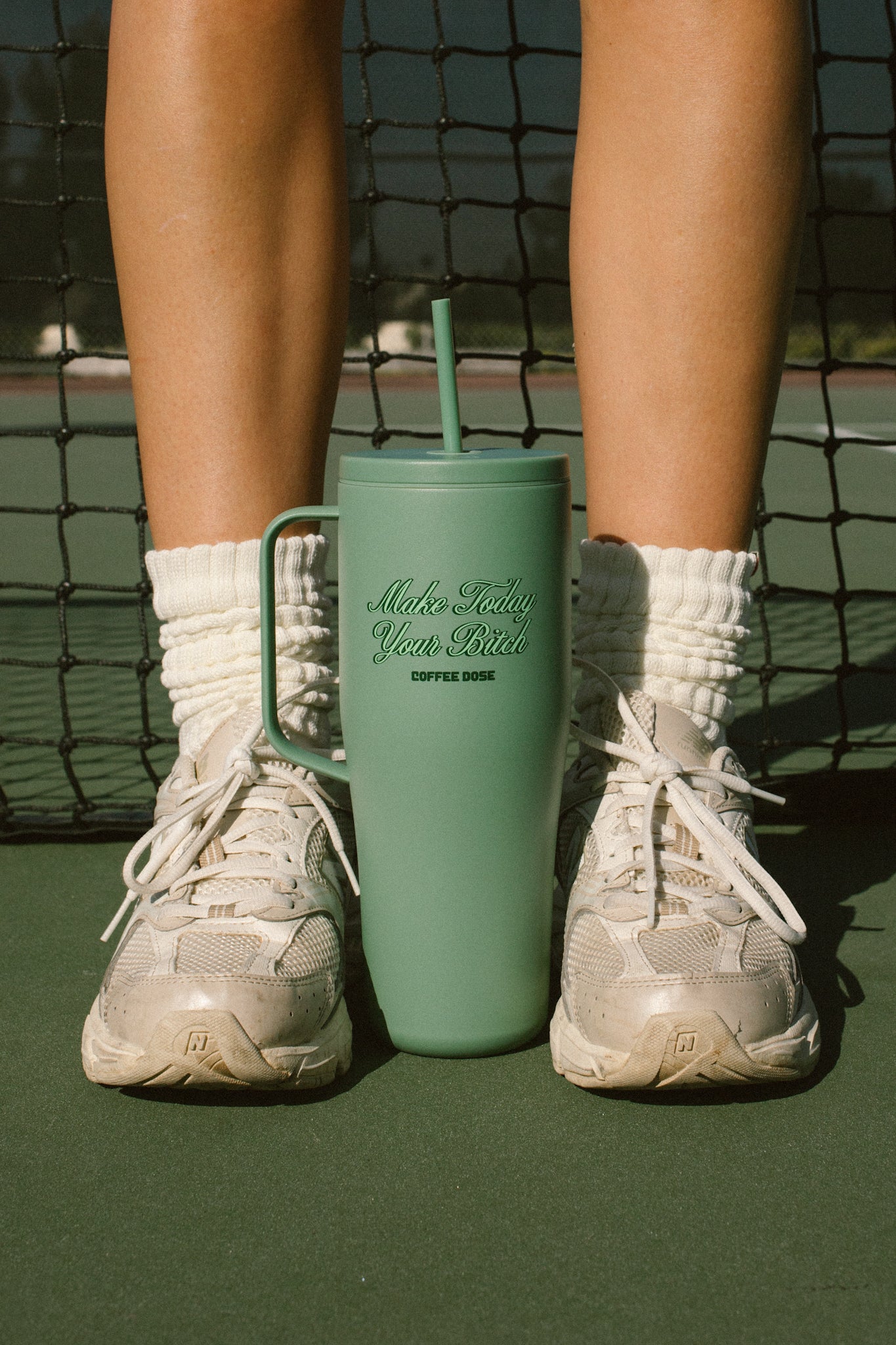 Person wearing white sneakers with a green tumbler that says make today your bitch on a tennis court