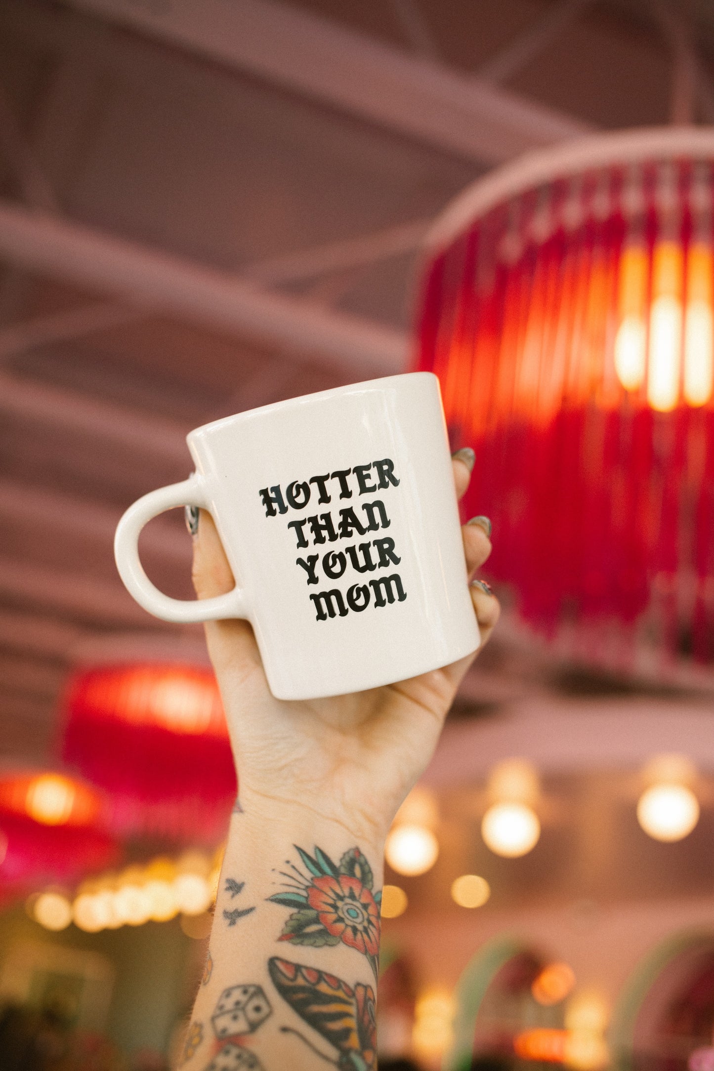 Hand holding a mug with 'HOTTER THAN YOUR MOM' text in a pink coffee shop