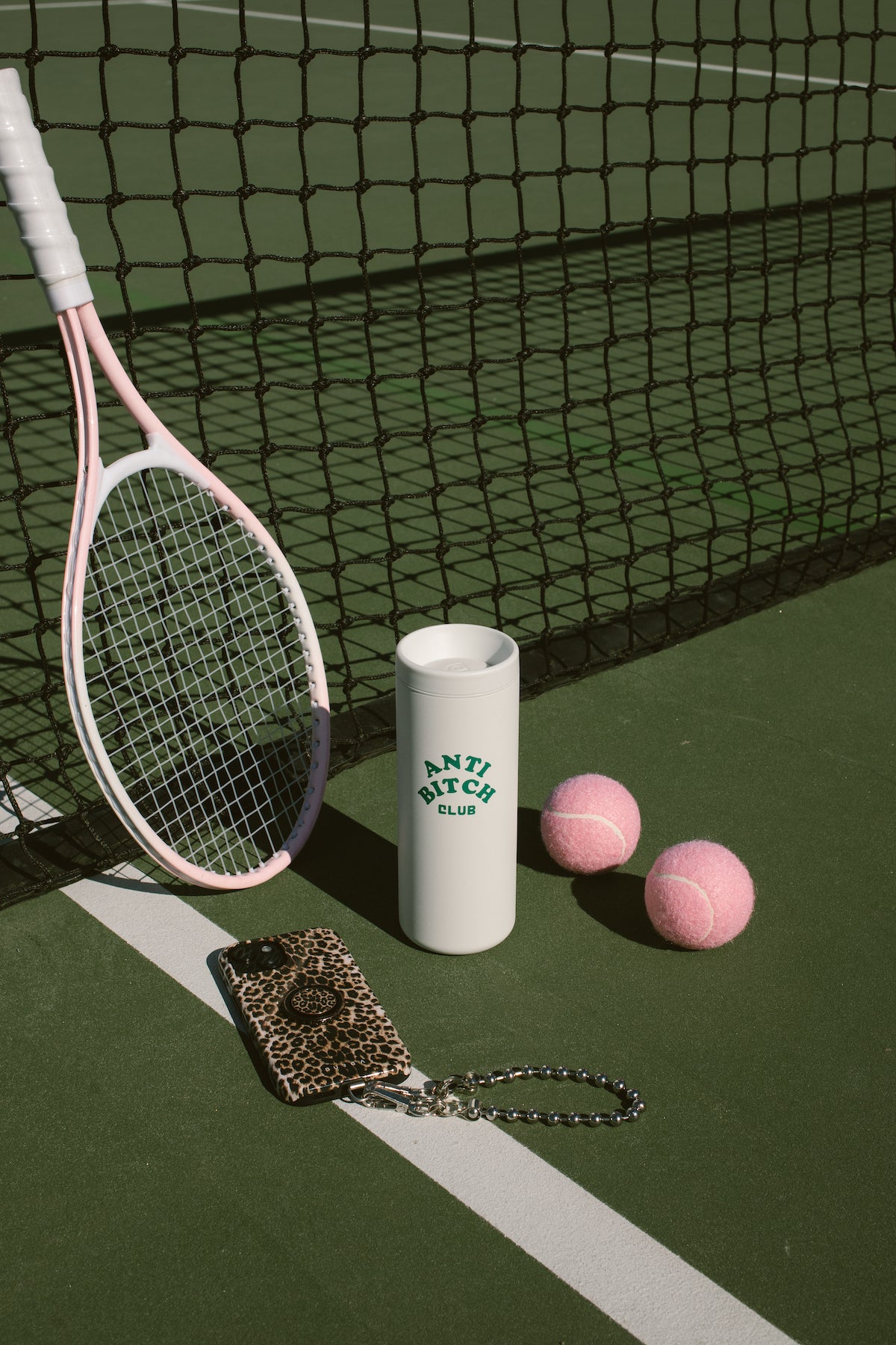 Pink tennis racket, white cup with 'Anti Bitch Club' text on the front, and pink tennis balls on a tennis court beside a leopard print iphone.