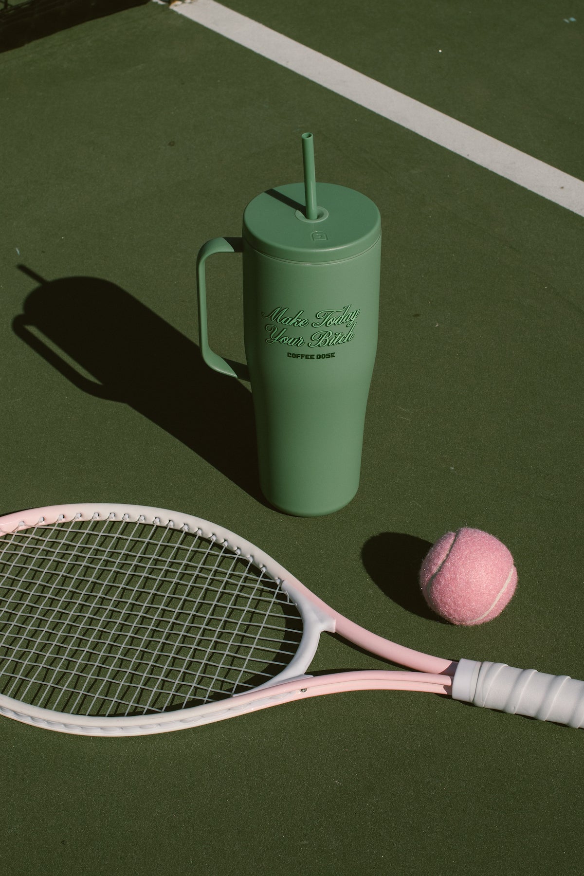 Green tumbler with a straw that says make today your bitch, tennis racket, and pink tennis ball on a green tennis court surface