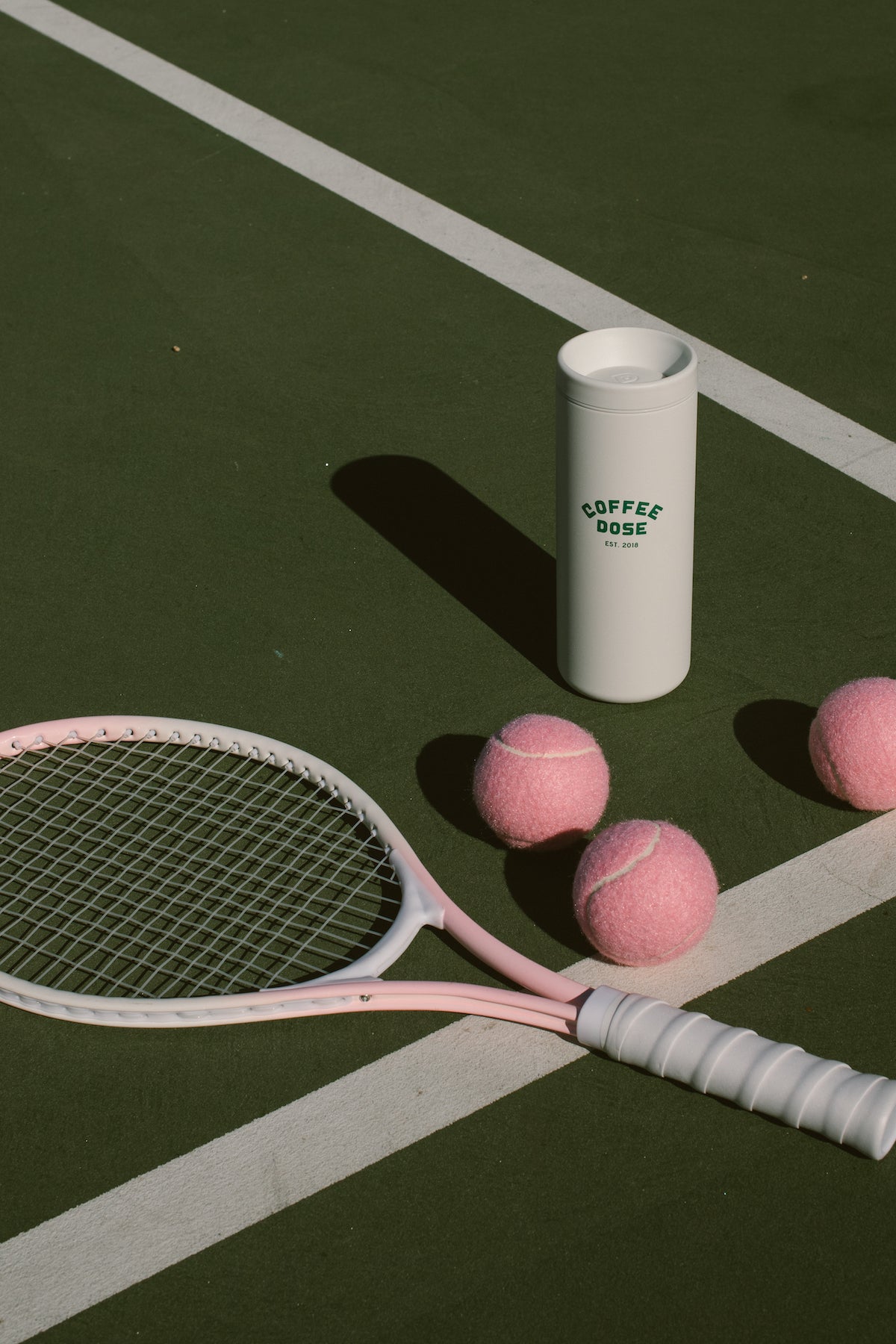 Tennis racket and pink tennis balls on a tennis court with a 'Coffee Dose' white tumbler.