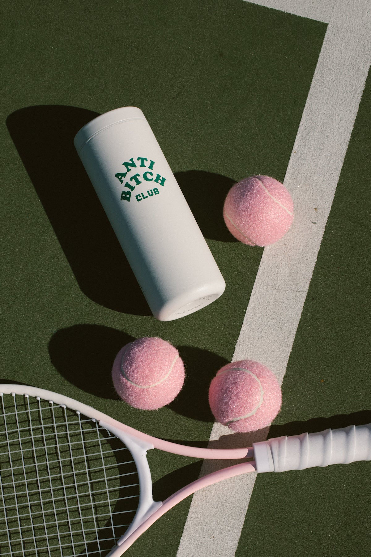 Tennis racket with pink tennis balls and a white tumbler labeled 'Anti Bitch Club' on a tennis court.