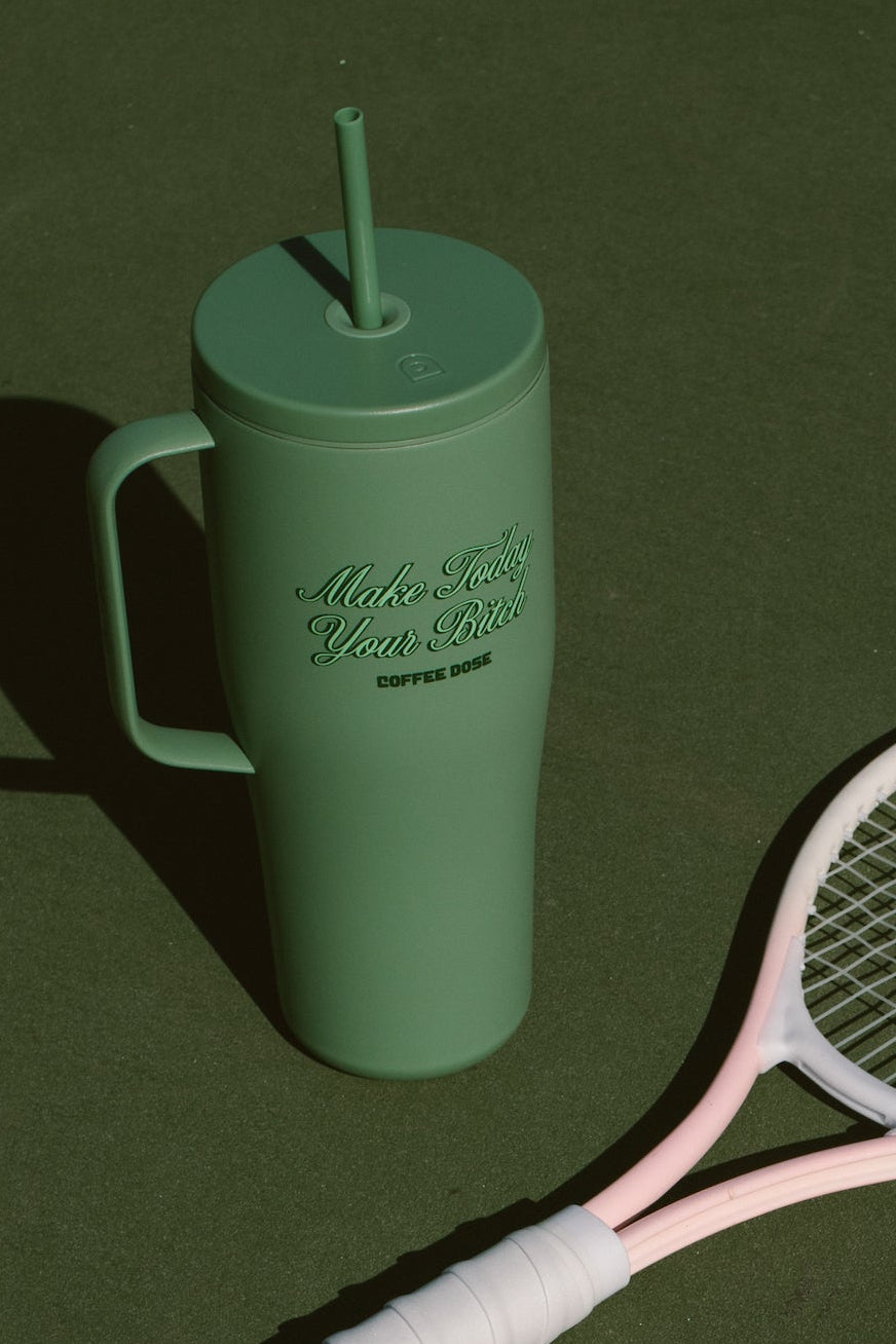 Green tumbler with a straw that says make today your bitch and pink tennis racket on a green tennis court surface