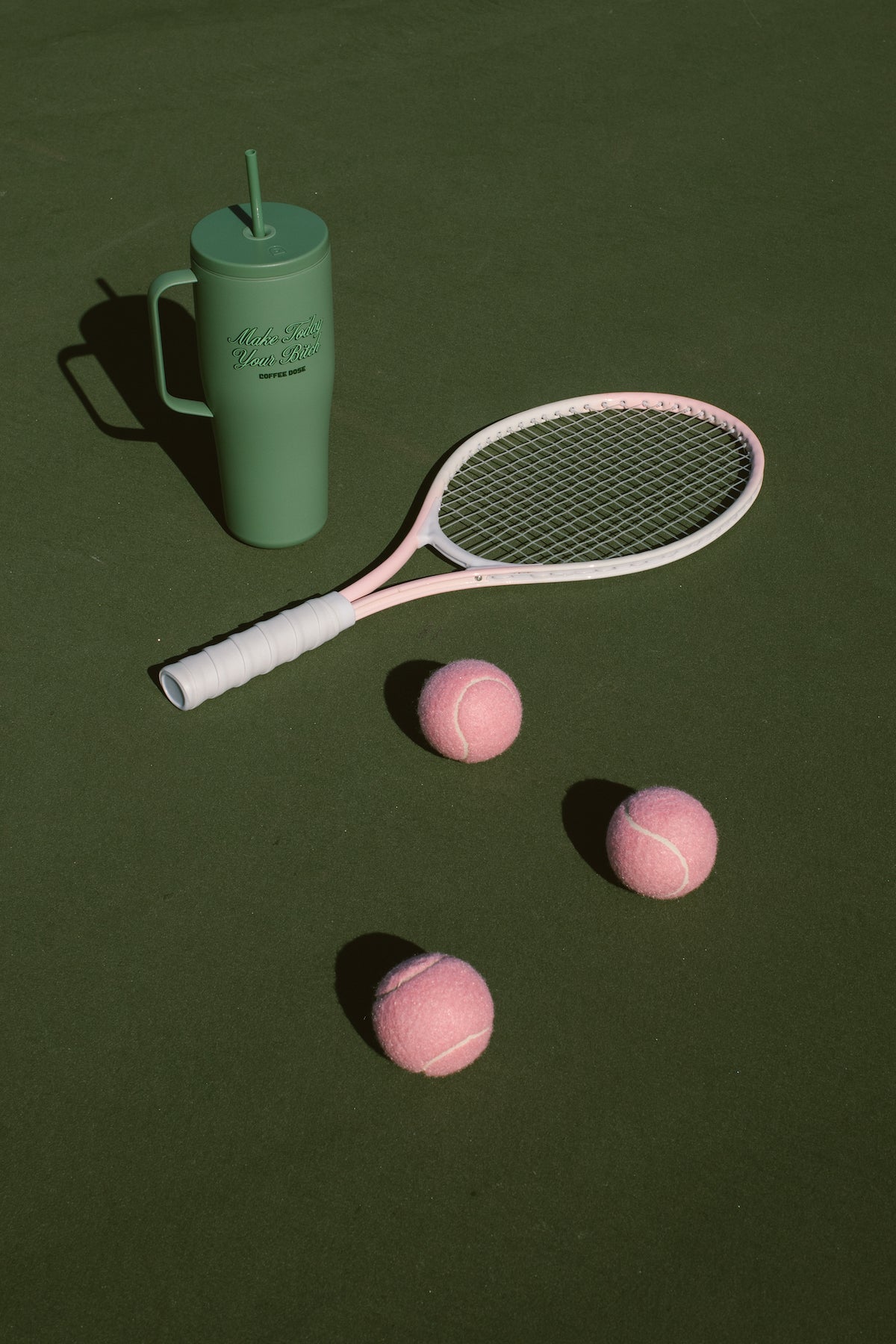 Green tumbler with a straw that says make today your bitch, tennis racket, and pink tennis balls on a green tennis court surface