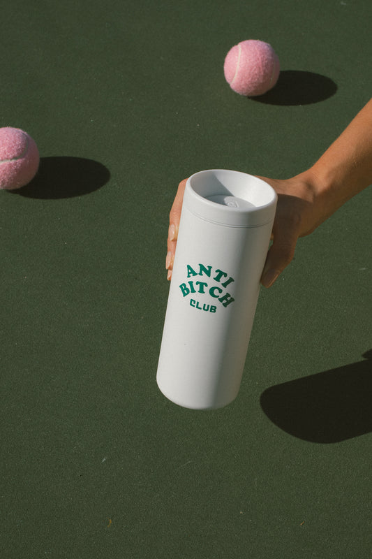 White tumbler with 'ANTI BITCH CLUB' text held by a hand, with pink tennis balls on a green background