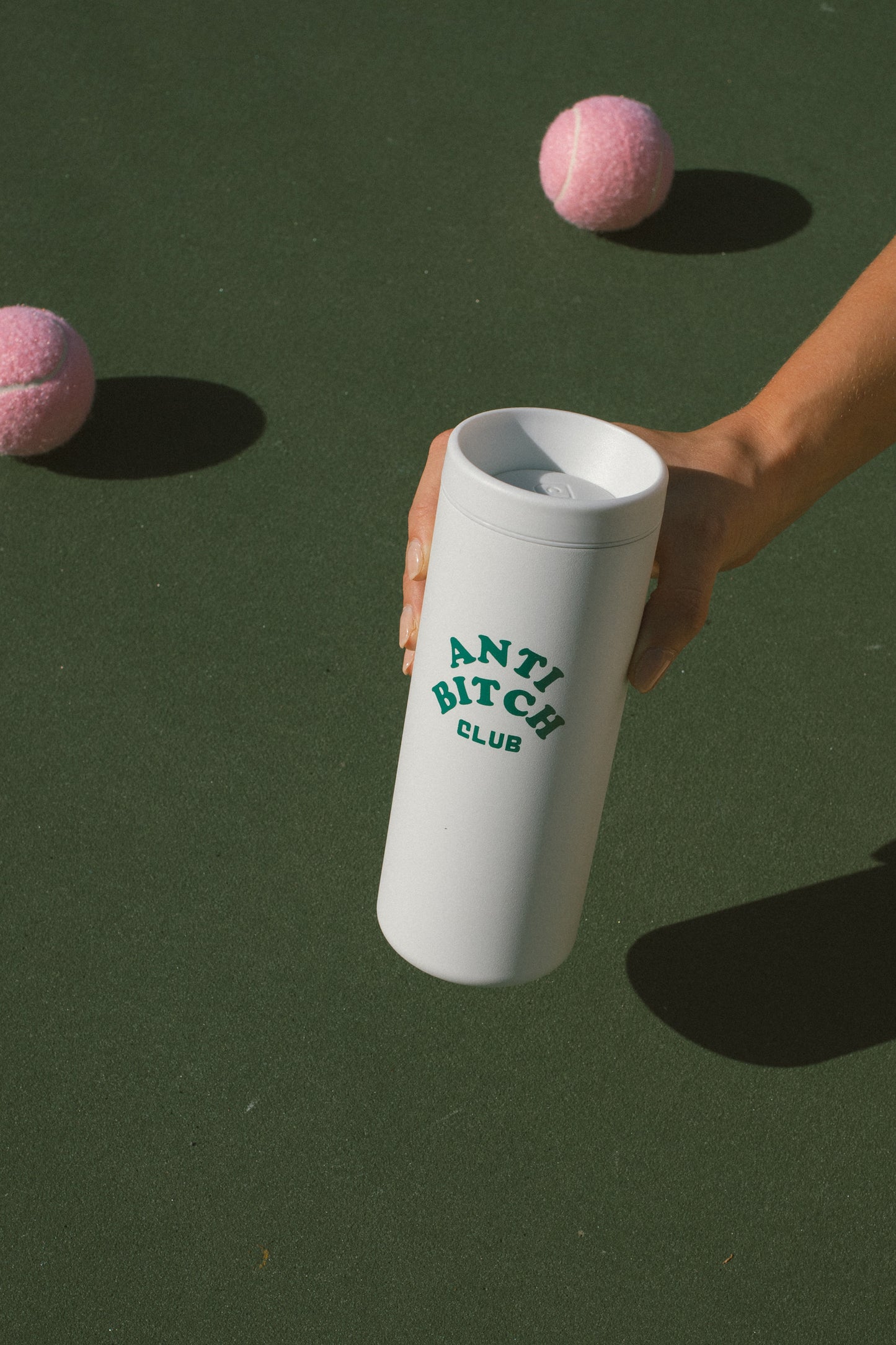 White tumbler with 'ANTI BITCH CLUB' text held by a hand, with pink tennis balls on a green background