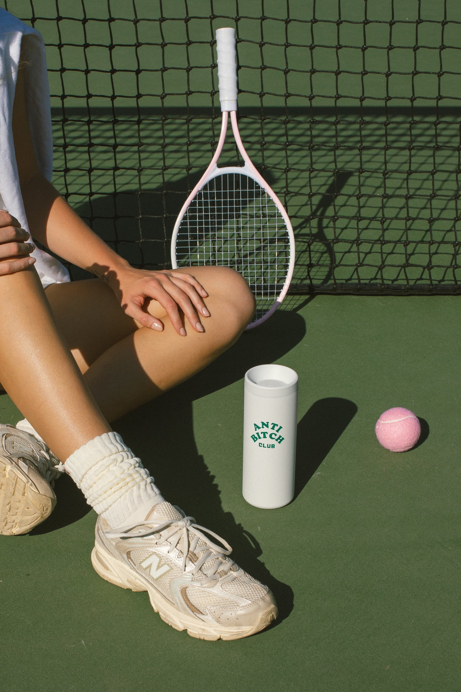 Person sitting on a tennis court with a tennis racket, white tumbler that says anti bitch club, and pink tennis ball.
