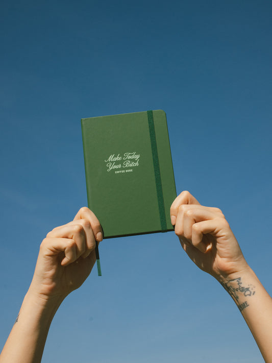 Green notebook that says 'make today your bitch' held up against a clear blue sky