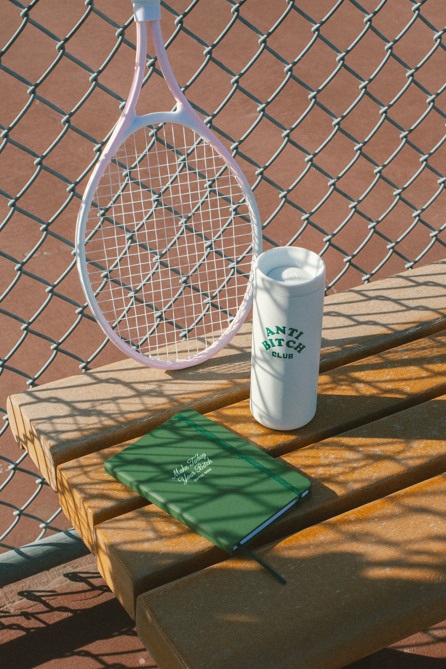 Tennis racket, green notebook that says 'make today your bitch', and white tumbler that says 'anti bitch club' on a wooden bench with chain-link fence in the background.